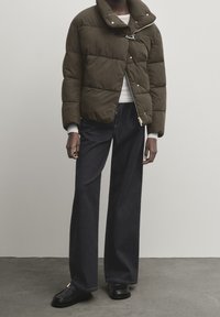 Olijfgroene pufferjack met horizontale stiksels, hoge kraag en een rits. Gecombineerd met donkerblauwe wijde jeans en zwarte schoenen.