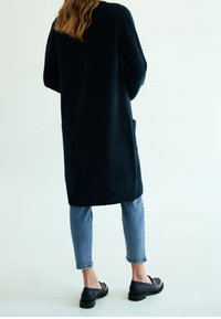 Lång marinblå cardigan av strukturerat tyg, med ribbade muddar, sidofickor och kombinerad med tajta jeans och svarta skor.