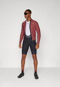 Muž oblečený v tmavě červené cyklistické bundě, bílé spodní vrstvě, černých cyklistických šortkách s laclem, černých cyklistických botách a velkých černých slunečních brýlích stojící na jednobarevném pozadí.