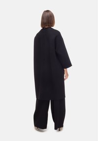 Cappotto nero oversize con taglio dritto, spalle cadenti e design minimalista. Texture liscia e linee pulite, indossato sopra pantaloni neri.