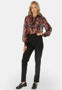 Przezroczysta czarna bluzka z kolorowym wzorem paisley, zawiązywanym dekoltem i długimi bufiastymi rękawami, w zestawieniu z czarnymi spodniami z wysokim stanem i błyszczącymi butami.