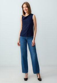 Top senza maniche di colore blu navy con scollatura drappeggiata e accessori decorativi in argento, abbinato a jeans a gamba larga blu e scarpe con tacco scuro.