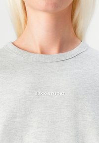 Sivá bavlnená tričko s okrúhlym výstrihom. Predná strana obsahuje vystúpený biely text "JJXX STUDIO" s hladkou textúrou a jemnými vizuálnymi detailmi.