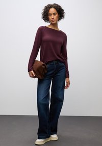 Bordeaux langemouwen top met ribbelstructuur, gecombineerd met donkerblauwe wijde jeans. Houdt een bruine suède clutch vast. Crèmekleurige sneakers.