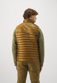 Brown puffer vest with horizontal quilting, high collar, and a shiny texture, worn over a green long-sleeve shirt and brown pants.