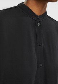 Camisa negra de botones con cuello alzado, que presenta una textura suave y pequeños botones negros a lo largo de la tapeta frontal.