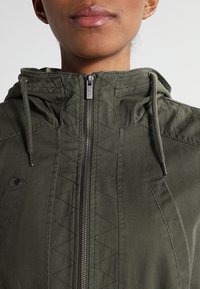 Olivgrön huvjacka tillverkad av tyg med en strukturerad finish. Funktioner inkluderar en framzipper, sidofickor och ett diamantquiltat mönster på axlarna.