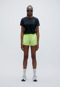 Αθλητική εμφάνιση που περιλαμβάνει μαύρο κοντομάνικο μπλουζάκι και φωτεινά λεμονί πράσινα σορτς. Τα αξεσουάρ περιλαμβάνουν αντανακλαστικά γυαλιά ηλίου και λευκά αθλητικά παπούτσια.