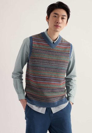 Jeune homme portant une chemise bleue à rayures sous un gilet tricoté à motifs multicolores et un pantalon bleu, debout les mains dans les poches.