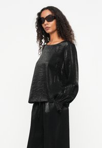 Čierny, lesklý top s dlhým rukávom a okrúhlym výstrihom, vo voľnom strihu. Obsahuje textúrovanú látku a zberané manžety pre dodatočný detail. Nosia sa slnečné okuliare.