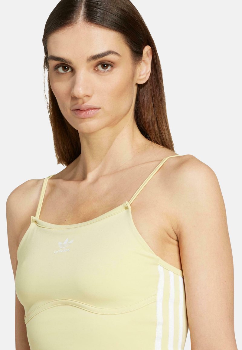 Femme avec des cheveux bruns lisses portant un débardeur Adidas jaune pâle à fines bretelles et trois bandes blanches sur le côté.