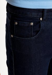 Jeans em denim azul escuro com uma textura suave, apresentando um pequeno bolso frontal e detalhes em botão metálico. Vista em close do tecido e do design.