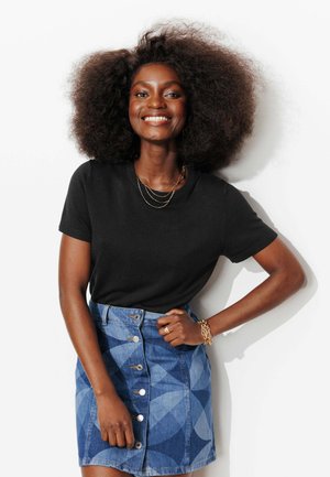 Femme souriante avec une coiffure afro naturelle, portant un t-shirt noir à manches courtes et une jupe en denim à motifs bleus avec boutons sur le devant, sur fond blanc.