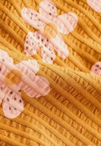 Tissu orange texturé avec un motif côtelé et de grandes fleurs roses présentant des taches foncées sur les pétales, montré en vue rapprochée.