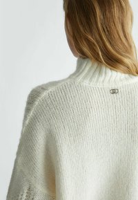 Maglione lavorato a maglia color crema con colletto alto a costine, motivo testurizzato e un accento metallico sottile del logo sul retro.