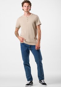 Beige Kurzarm-Henley-Shirt mit drei Knöpfen, kombiniert mit blauen Jeans und schwarzen Sneakers. Glatte Textur mit lockerer Passform.