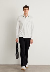 Hvit langermet polo-skjorte med kontrastgrønn stripe på kragen, parret med svarte bukser og hvite joggesko. Svart veske slengt over skulderen.