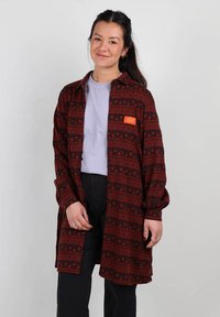 Veste à motifs rouges avec accents noirs, dotée d'une poche avant et d'une fermeture à bouton. En dessous, un t-shirt léger de couleur violet clair est visible.
