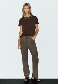 Young woman wearing a dark brown short-sleeve t-shirt, brown suede pants, and black open-toe slide sandals, standing against a plain white wall.