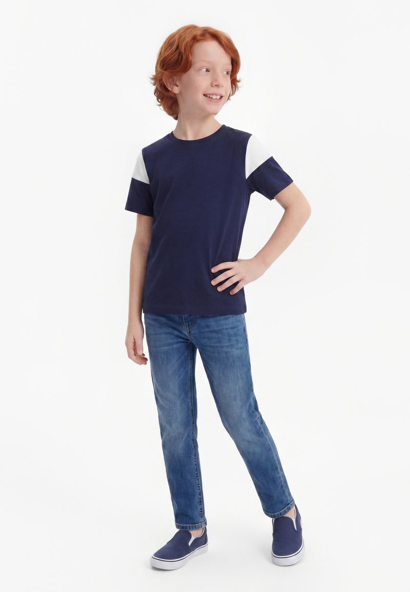 Enfant aux cheveux roux debout, souriant, main sur la hanche, portant un t-shirt bleu marine et blanc à manches courtes, un jean bleu et des chaussures bleu marine sans lacets.