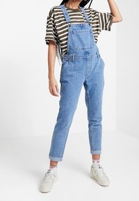 Salopette en denim bleu clair avec bretelles ajustables, poche avant et jambes fuselées, associée à un t-shirt rayé marron et blanc et des baskets blanches.