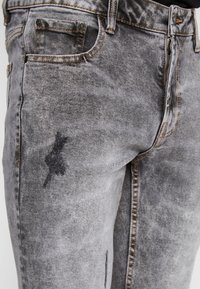 Grå denimjeans med en blekt tvätt, som har slitna områden och ett subtilt svart märke på låret, med kontrasterande sömnadsdetaljer.