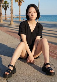 Ung kvinde i sort outfit, der sidder med benene over kors på en brolagt sti ved havet med palmer i baggrunden.