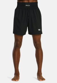 Atleet in zwarte sportshorts met elastische tailleband en witte logo's, die blote voeten op een witte achtergrond staat.