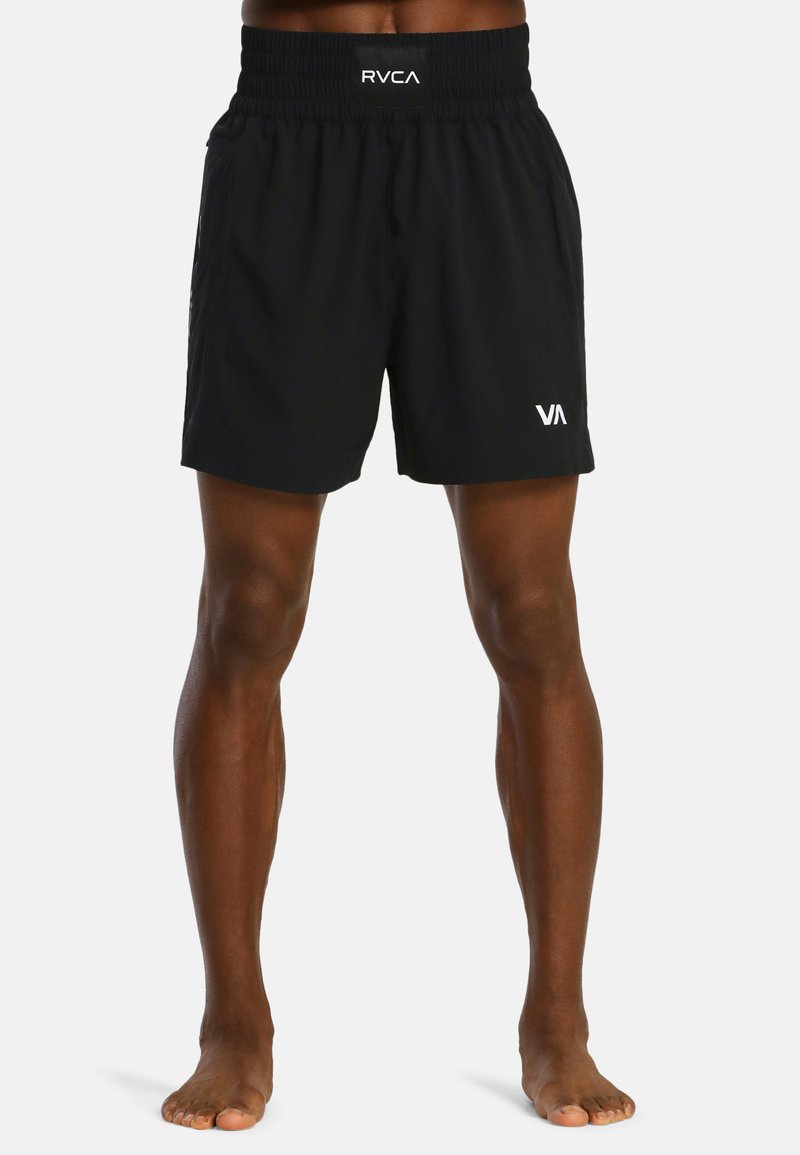Atleet in zwarte sportshorts met elastische tailleband en witte logo's, die blote voeten op een witte achtergrond staat.