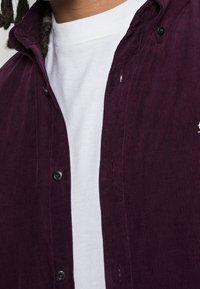 Camicia in velluto a coste viola con bottoni neri, indossata sopra una t-shirt bianca a girocollo. Righe testurizzate visibili sul tessuto.