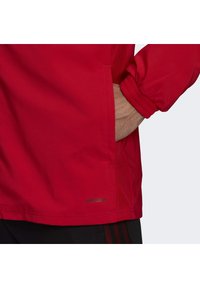 Chaqueta roja con puños elásticos y bolsillo lateral, con tejido suave y un logotipo "AEROREADY" visible cerca del dobladillo.