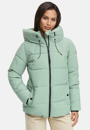 Femme portant une doudoune matelassée vert clair avec un col montant et une fermeture éclair noire, se tenant avec une main dans la poche.