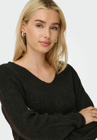 Maglione nero a maglia con scollo a V e maniche lunghe e voluminose. Il modello indossa orecchini a cerchio d'argento. Il tono della pelle è neutro; i capelli sono lunghi e biondi.