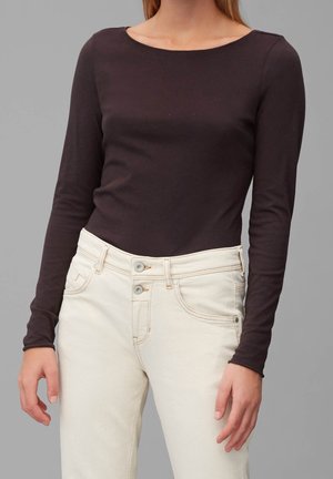 Frau trägt ein figurbetontes dunkelbraunes Langarmshirt und hoch taillierte, elfenbeinfarbene Hosen mit doppelten Knöpfen vor einem schlichten grauen Hintergrund.