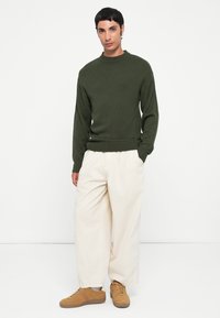 Donkergroen gebreid vest met een ronde hals, gecombineerd met losse beige broek en tan sneakers. De stof lijkt zacht en textuur te hebben.