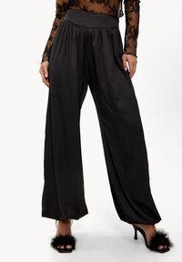 Pantalon large en satin noir à la texture lisse, taille haute avec un design plissé. Associé à des sandales à talons noires ornées d'accents en fourrure.