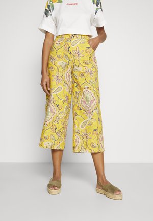Femme portant un pantalon jaune taille haute et jambes larges avec un imprimé floral cachemire, une chemise blanche à manches courtes et des sandales plateformes vert olive.
