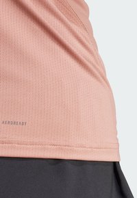 Ljusrosa träningstop med en strukturerad, fukttransporterande tyg och "AEROREADY"-logotyp, i kombination med en svart kjol nedanför.