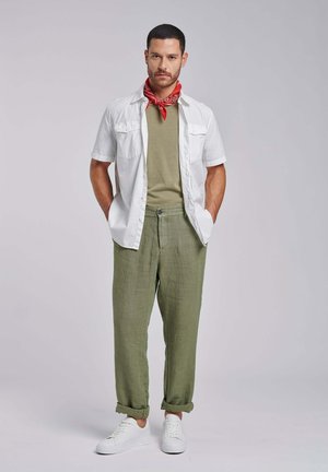 Mann steht mit den Händen in den Taschen, trägt ein weißes kurzärmliges Hemd, olivgrüne Hose, grünes T-Shirt, roten Halstuch und weiße Sneaker.