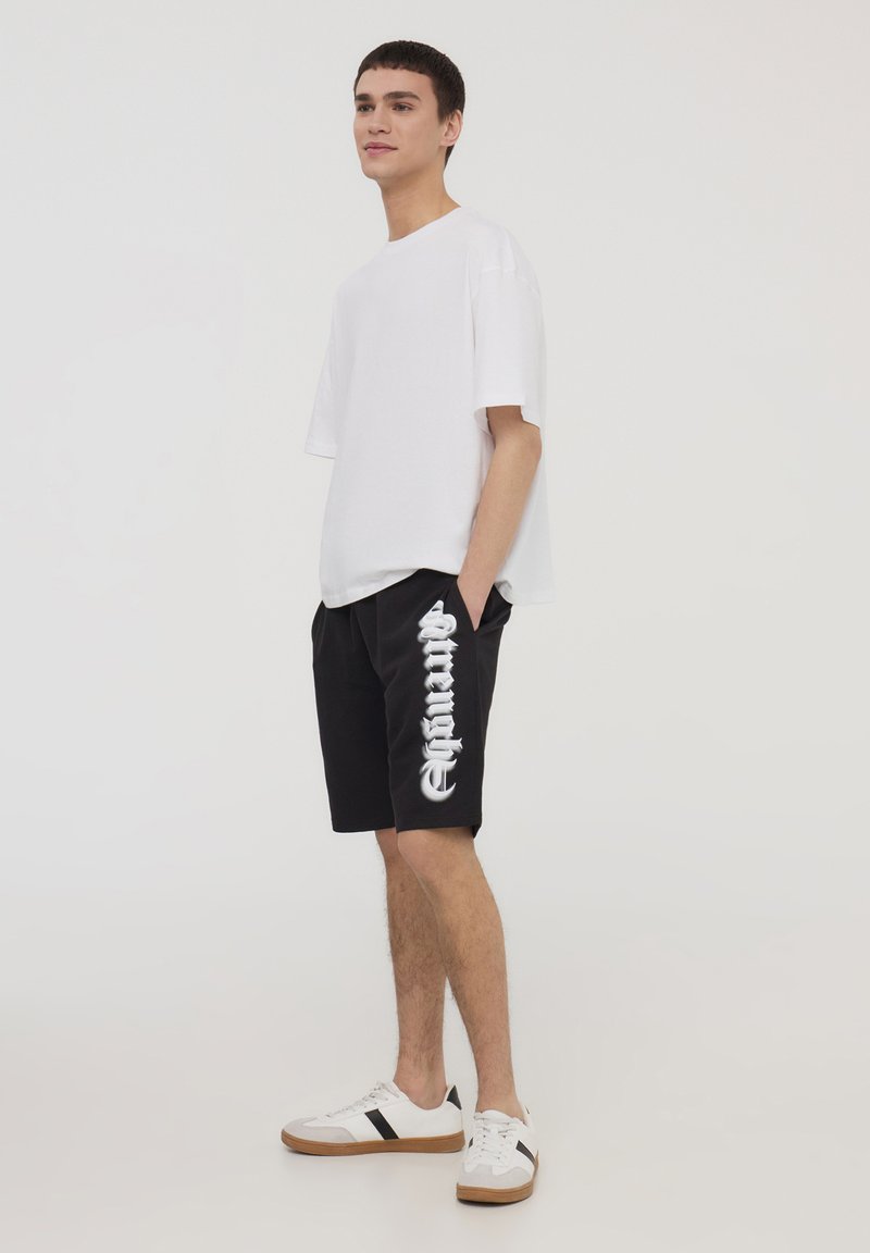 T-shirt bianco oversized abbinato a pantaloni corti neri con testo bianco. Il modello indossa sneakers bianche con dettagli marroni. Abbigliamento semplice e casual.