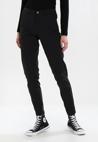 Černé slim-fit kalhoty s hladkou texturou, vybavené zapínáním na knoflík a kapsami. Nošeny s černými teniskami s vysokým svrškem.