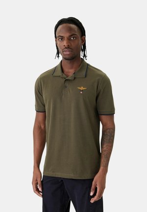 Giovane con capelli intrecciati che indossa una polo verde oliva con un emblema ricamato e pantaloni scuri, in piedi davanti a uno sfondo semplice.