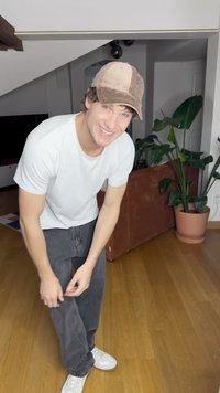 Witte t-shirt, grijze spijkerbroek en witte sneakers. De persoon draagt een bruine corduroy pet. Binnenomgeving met een plant en een bruine bank zichtbaar.