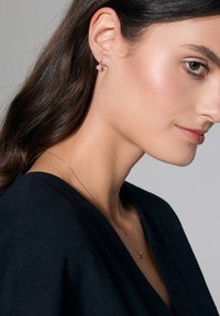 Złote kolczyki w kształcie hooop z kwiatowym wzorem, z różowym akcentem. Noszone przez modelkę o ciemnych włosach, ukazujące szczegóły i blask kolczyków.