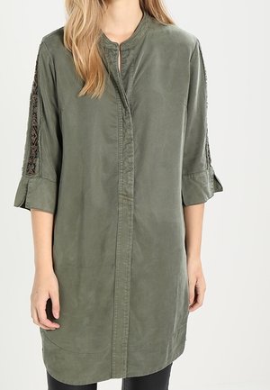 Femme portant une longue tunique vert olive avec des manches trois-quarts munies de panneaux brodés et de poignets repliés.