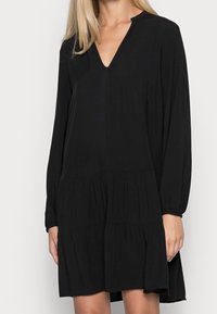 Vestido negro de manga larga con diseño de cuello en V, dobladillo en capas y tejido suave. Presenta un ajuste relajado y sutiles fruncidos en los hombros.