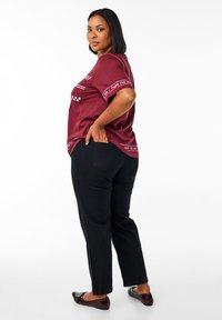Bordeaux t-shirt met contrasterende grafische details, gecombineerd met zwarte broek en glanzende zwarte schoenen. Model is van opzij weergegeven.