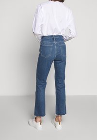 Jeans de denim en azul oscuro con una cintura alta, pierna recta y dobladillo deshilachado, combinados con una blusa blanca de manga larga y zapatillas blancas.