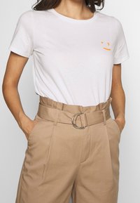 White cotton t-shirt with short sleeves and a small orange smiley face graphic on the chest, paired with beige high-waisted trousers with a belt.
