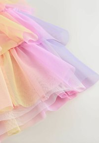 Couches de tissu transparent en jaune pastel, rose et violet réunies à la taille, créant un détail de jupe volantée et fluide.